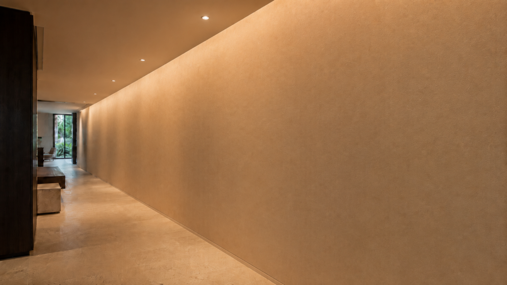 Long plaster corridor wall evenly illuminated from ceiling to floor by recessed wall-washing downlights.